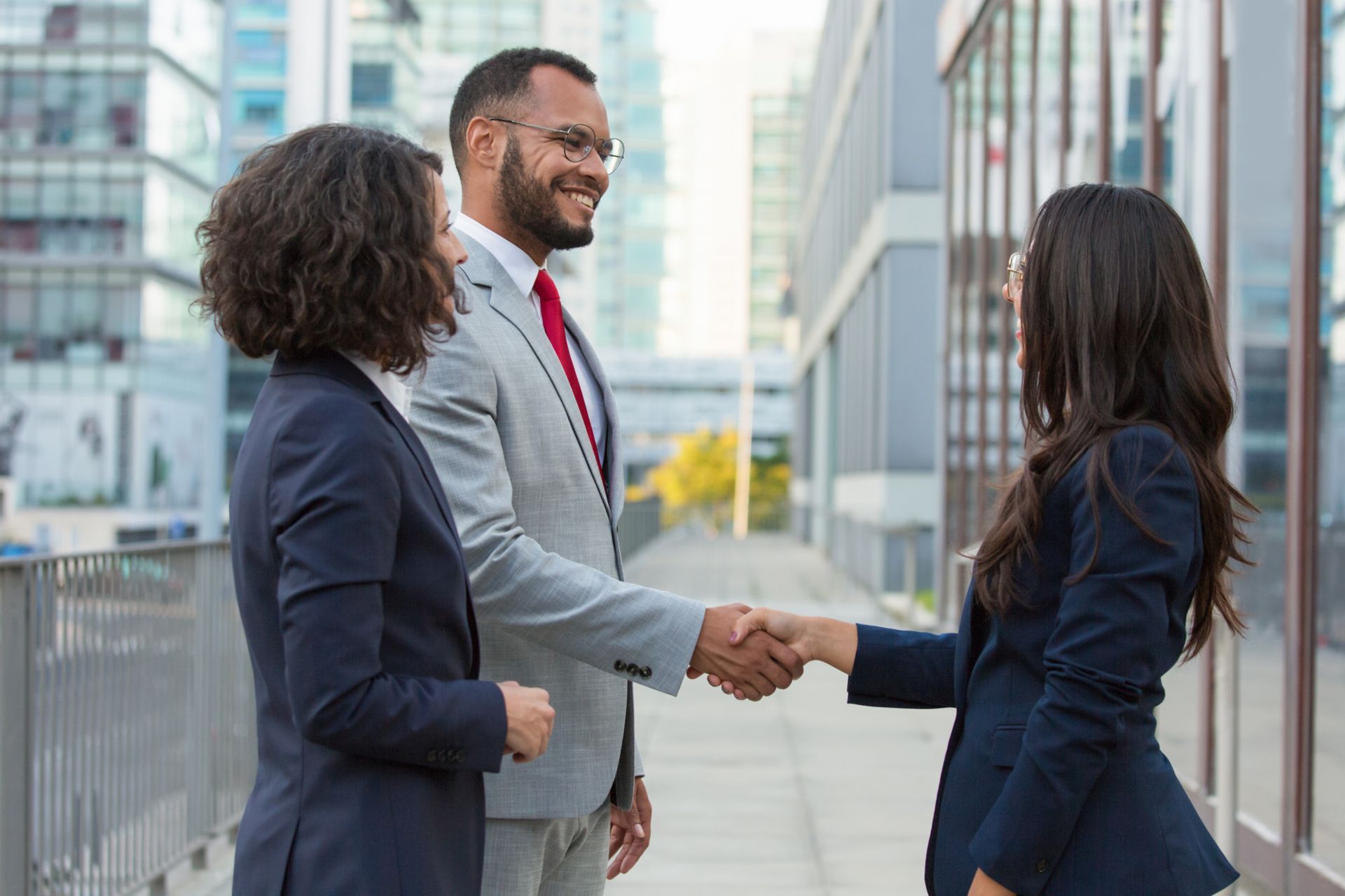 Businesspeople shaking hands outdoors, near buildings. Man smiles, two women watch.