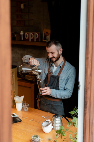 Barista pouring coffee from a silver pot into a glass; smiling. Inside a coffee shop, wooden counter, and plants.