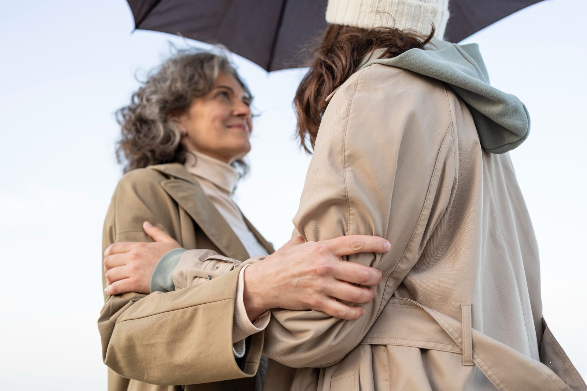 Woman in raincoat hugs another under an umbrella on a cloudy day.