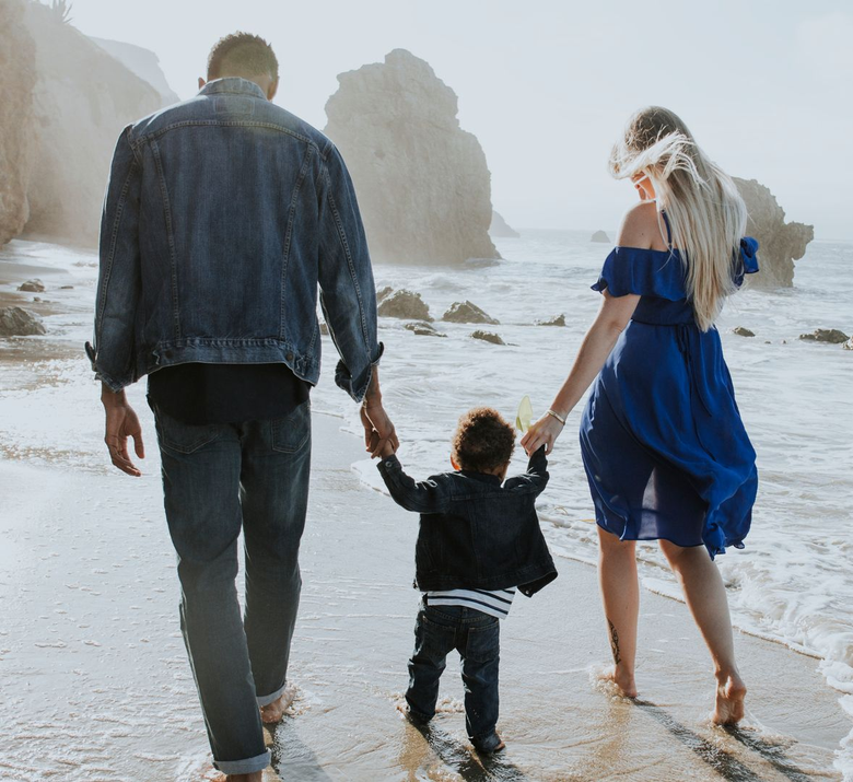Family walking on a beach; parents hold a child's hands; ocean in the background.