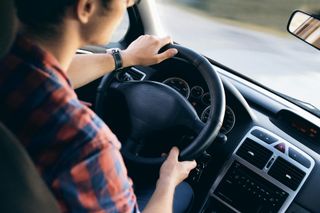 Person driving a car, hands on steering wheel, wearing a red plaid shirt.
