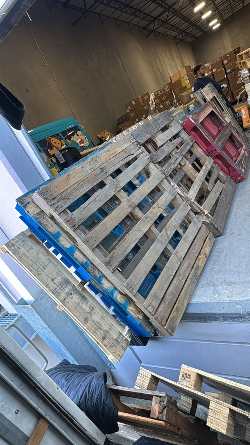 Stacked wooden pallets in a warehouse aisle, with blue and pink pallet edges visible.