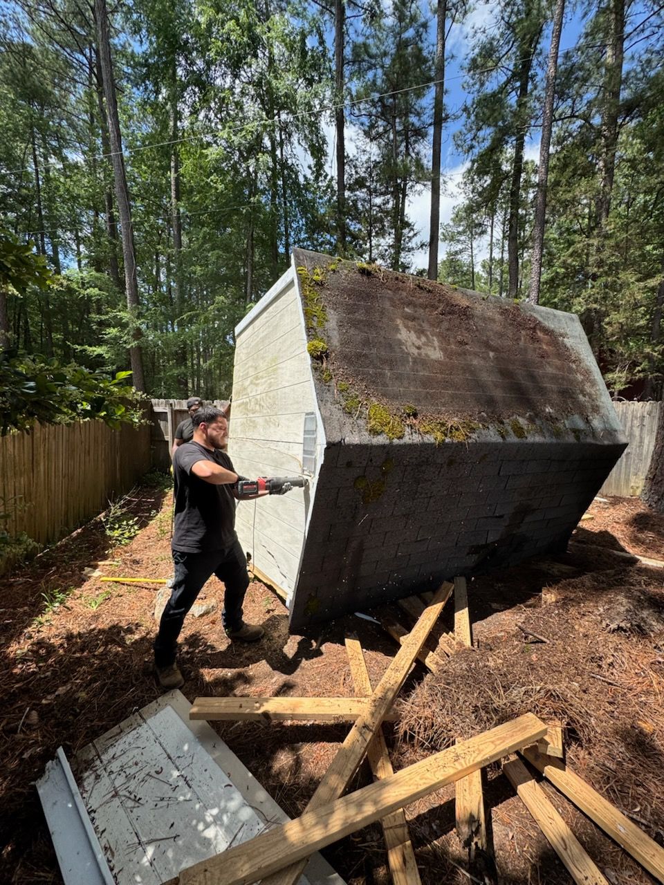 Person pushing a large concrete block in a wooded yard, with lumber and debris nearby.