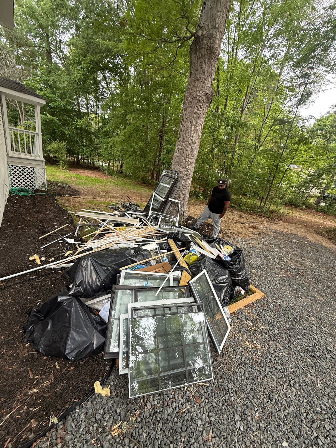 Piled storm debris and broken window frames beneath a tree beside a house, with a person standing nearby.