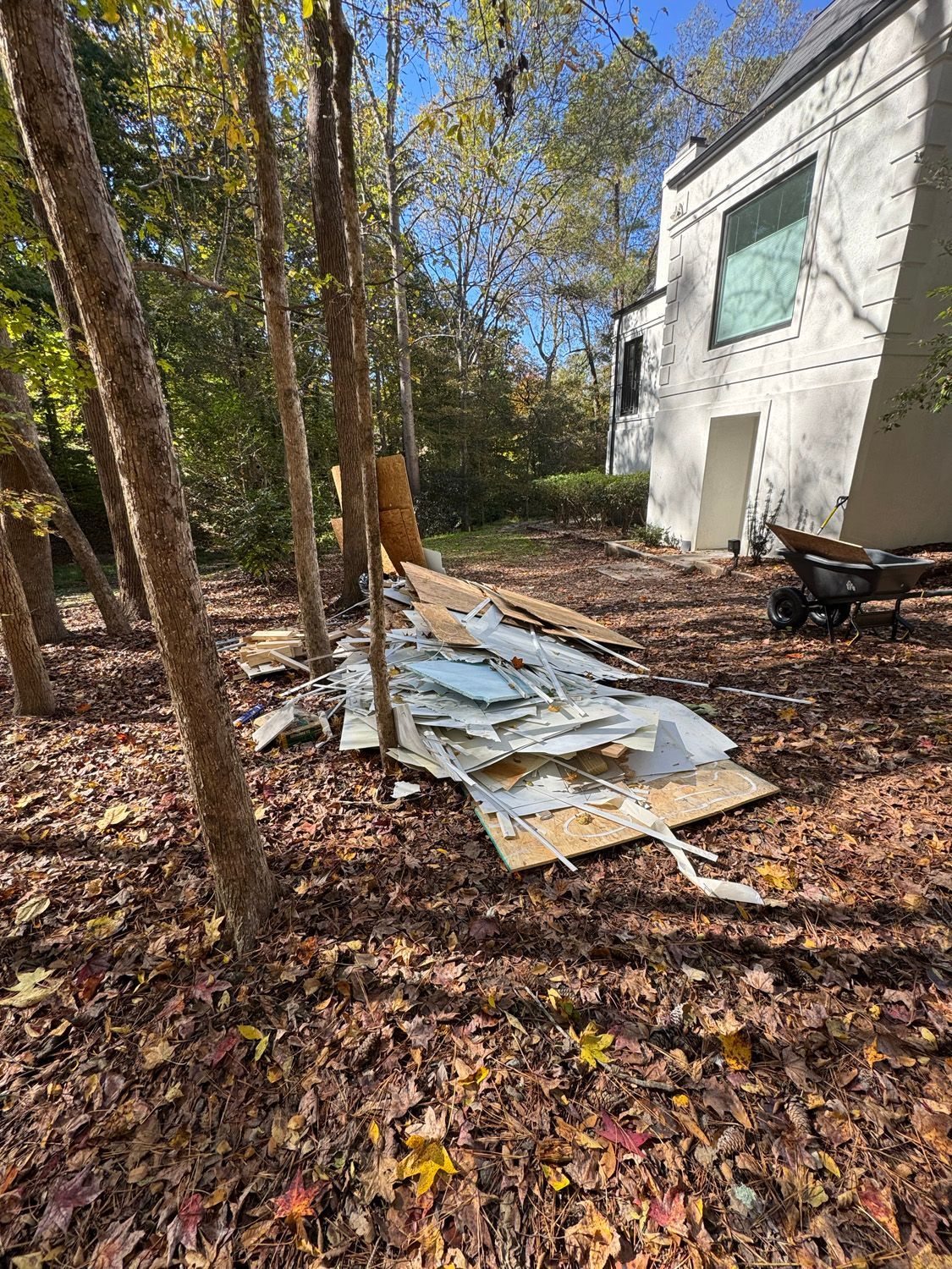 Outdoor debris pile beside a white house in a wooded yard, with leaves scattered on the ground.