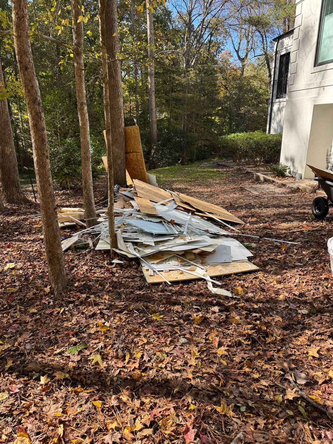 Pile of broken white panels and debris in a wooded yard beside a house