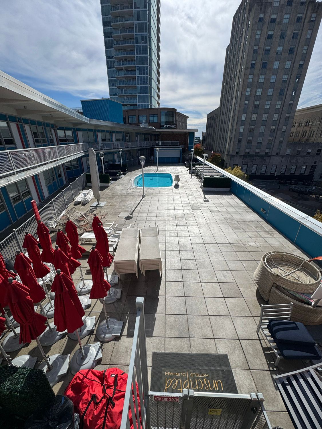 Rooftop pool deck with red umbrellas, lounge chairs, and city buildings under a blue sky