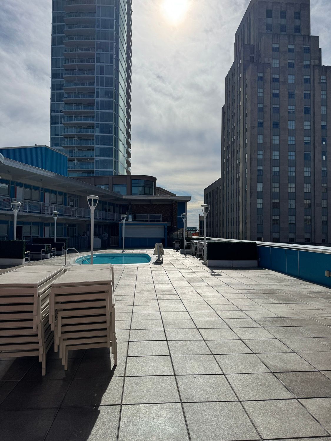 Rooftop terrace with pool, stacked lounge chairs, and city buildings under bright sun