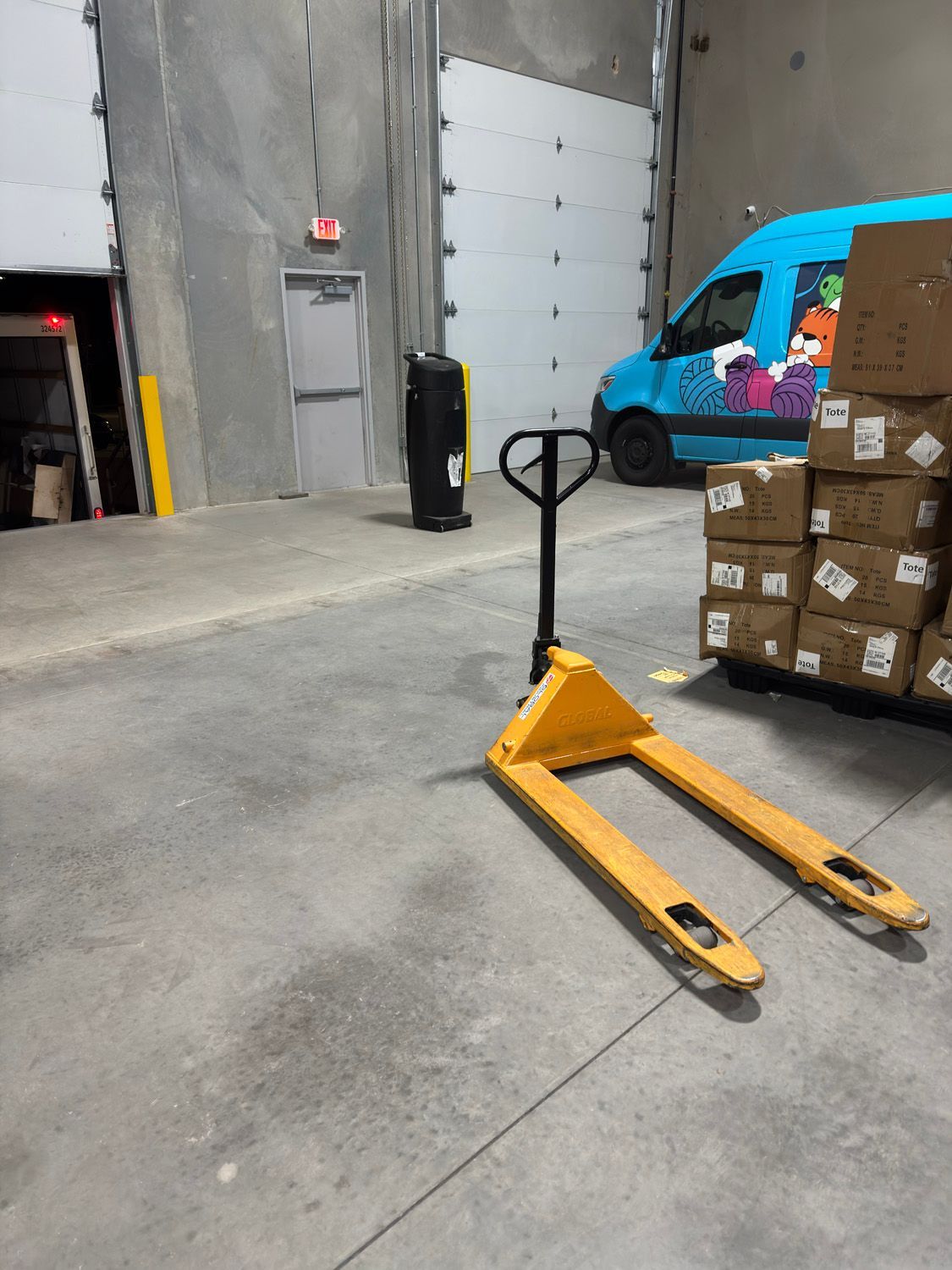 Yellow pallet jack in a warehouse beside stacked boxes and a blue van.