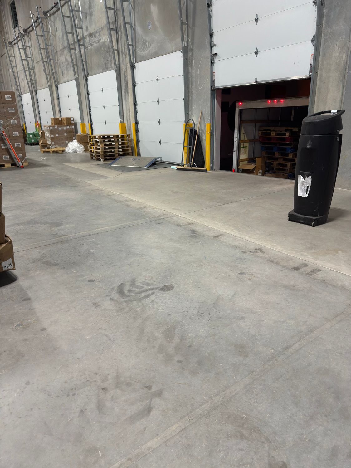 Warehouse loading dock with concrete floor, tall gray doors, stacked pallets, and a black trash bin.