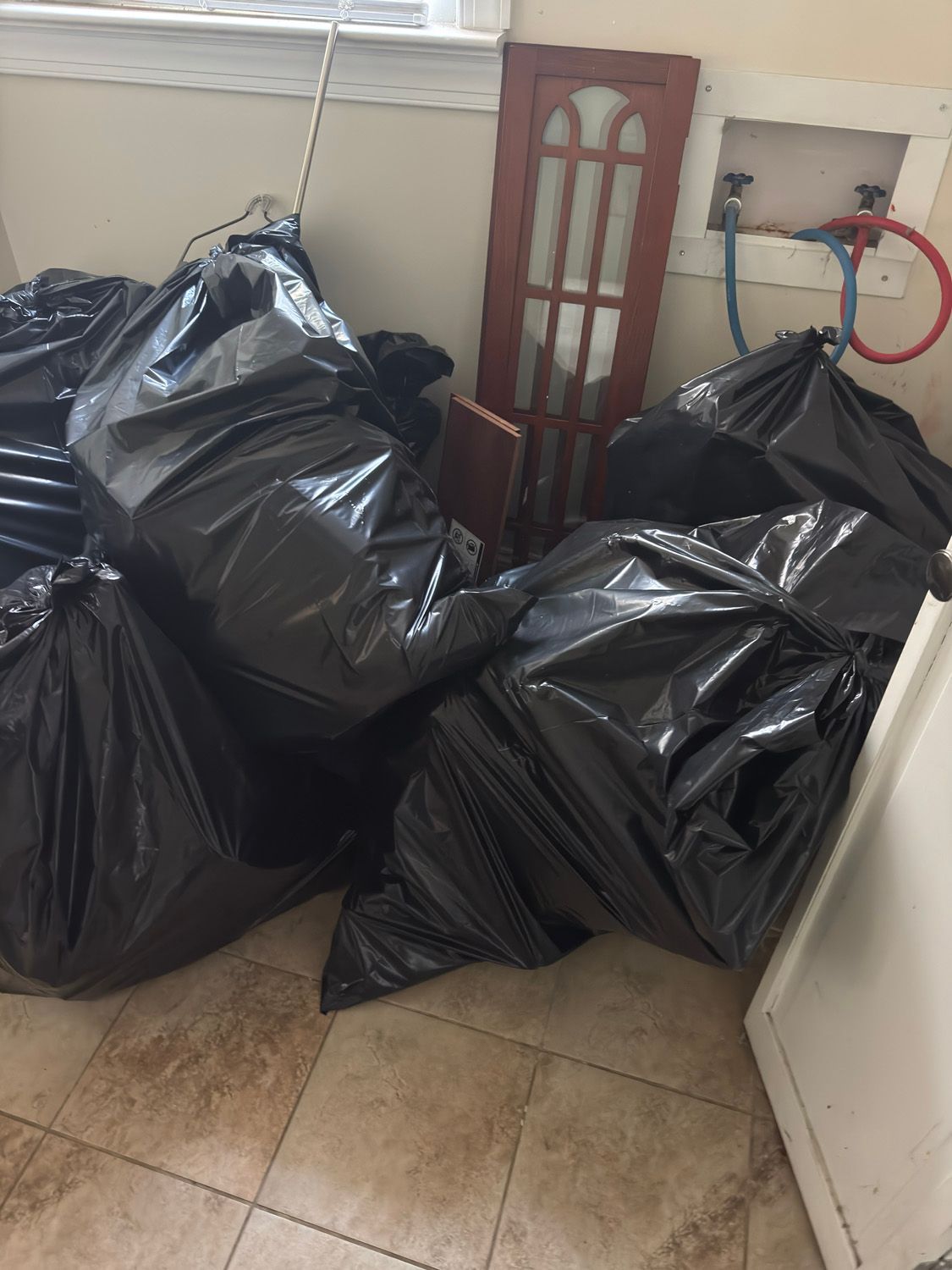 Black trash bags piled in a tiled room beside a cabinet and wall-mounted pipes.