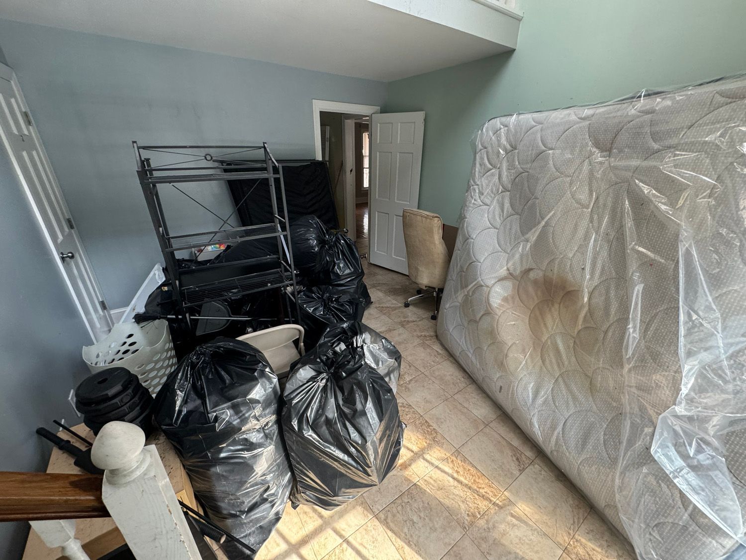Cluttered room with black trash bags, a metal rack, and a large plastic-covered item against the wall.