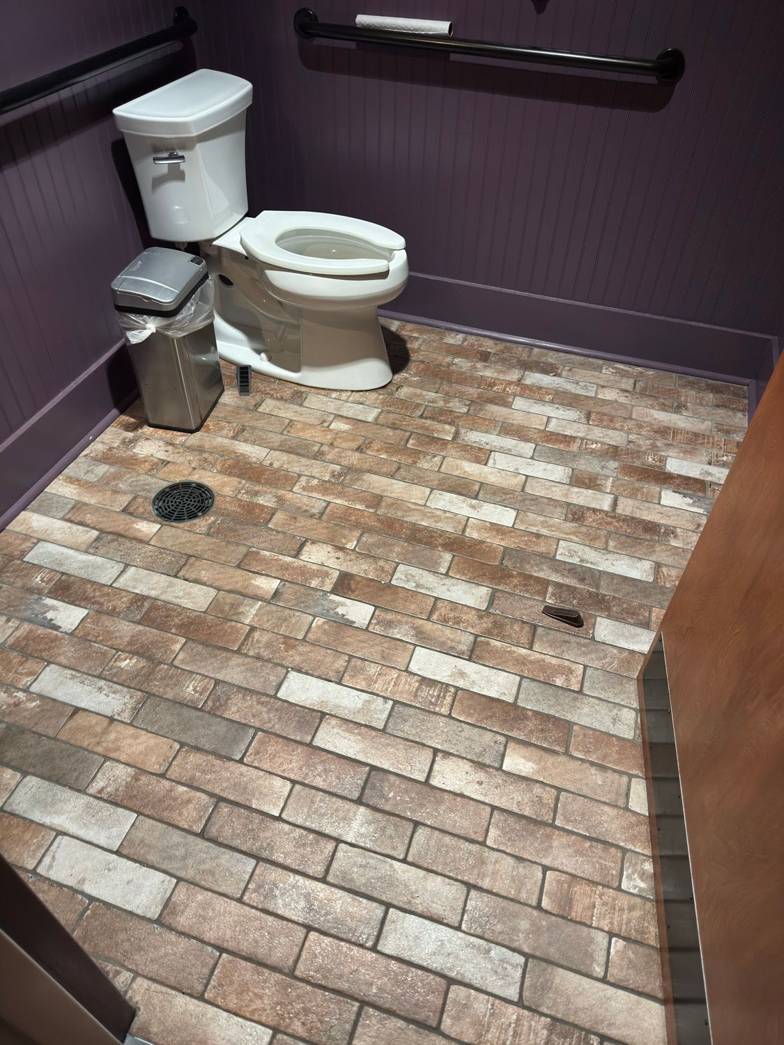 Small restroom with a toilet, trash can, and dark walls over a brick-pattern floor.