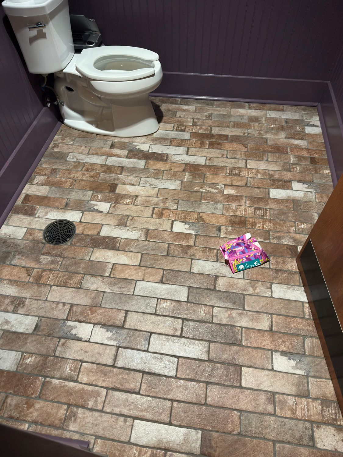 Small brick-patterned bathroom with a toilet, floor drain, and a colorful wrapper on the floor.