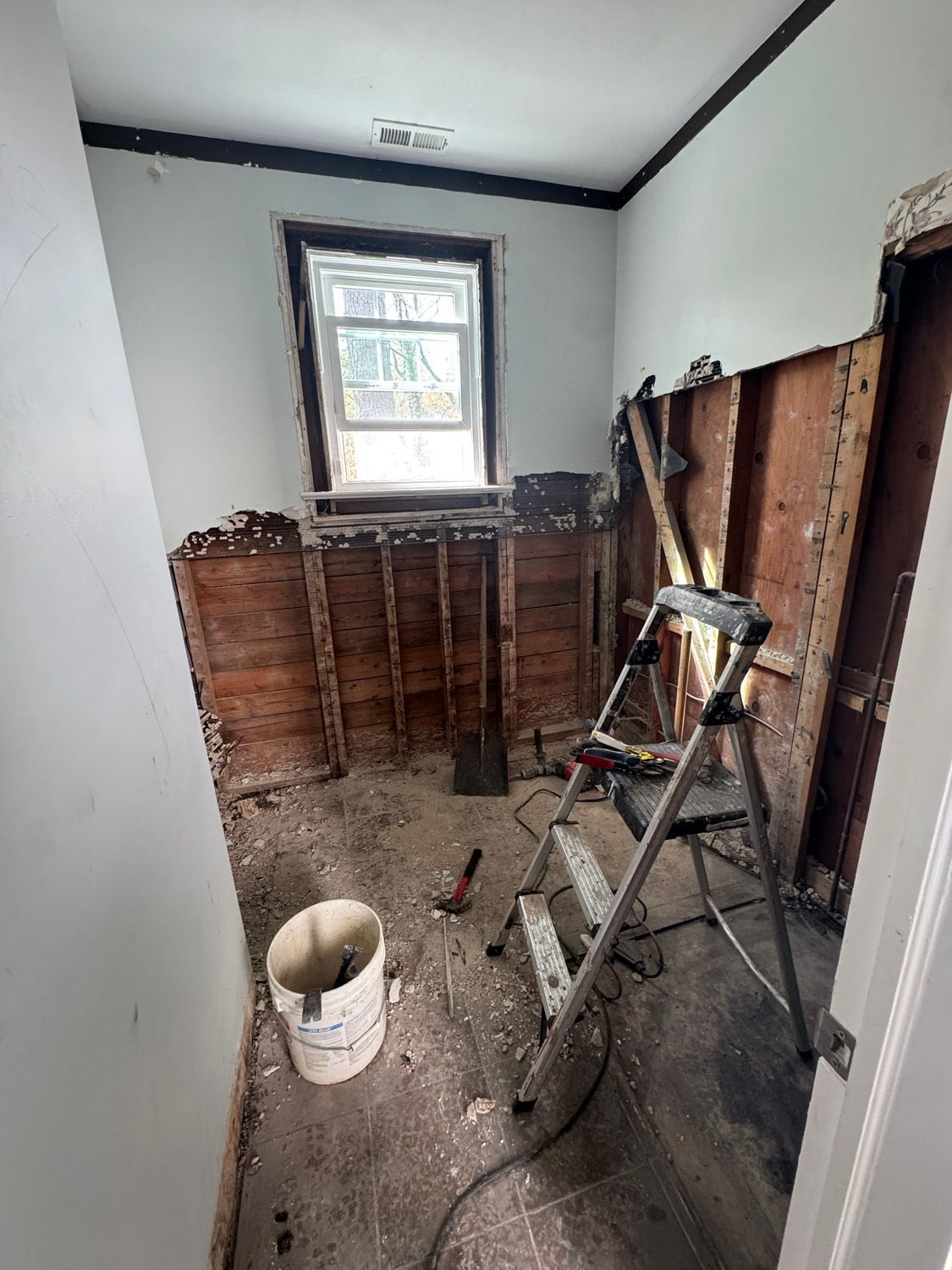 Small room under renovation with exposed wood walls, a ladder, bucket, and debris on the floor