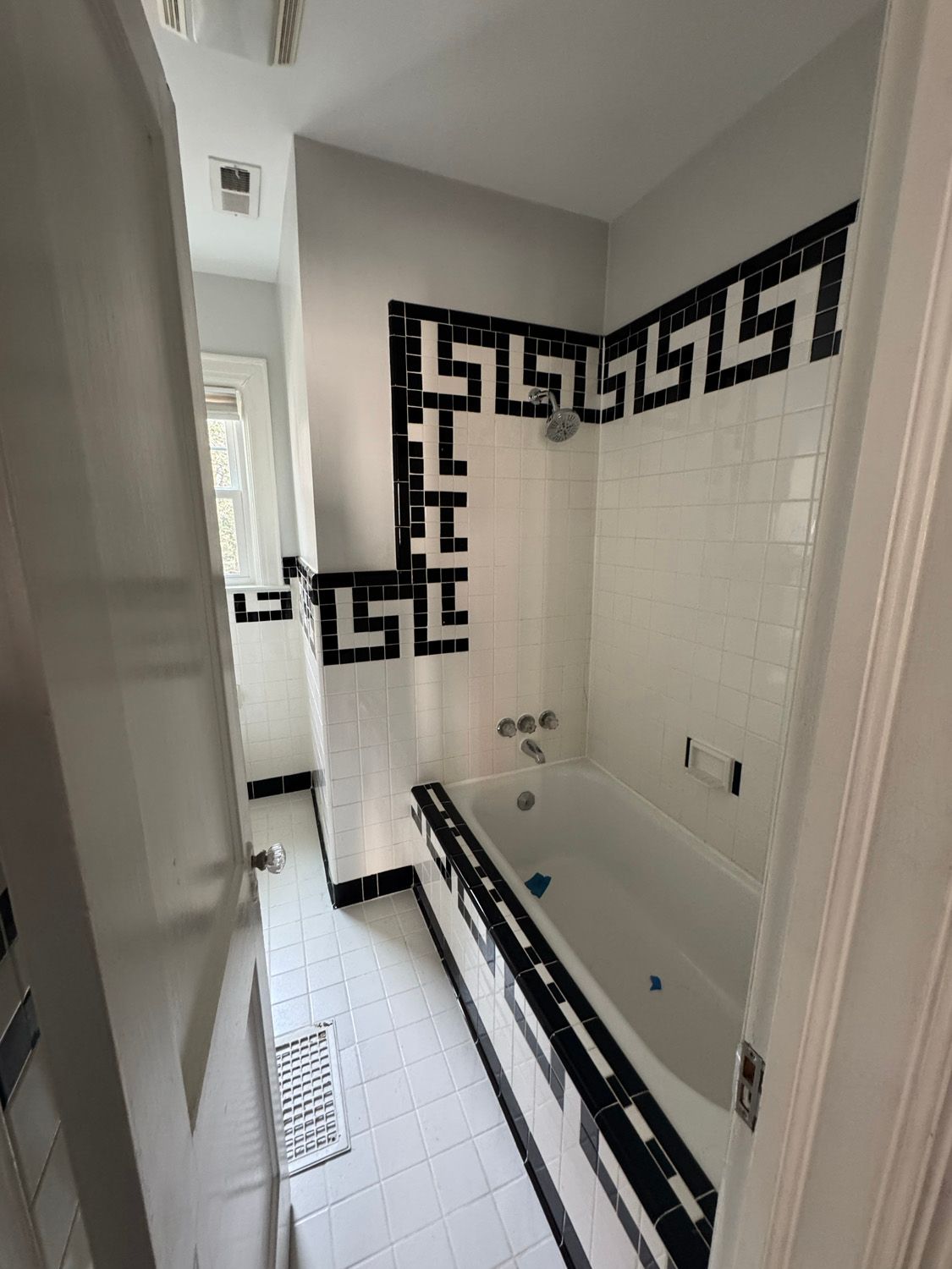 Narrow bathroom with a white tub, black Greek key tile border, and a window at the far end.
