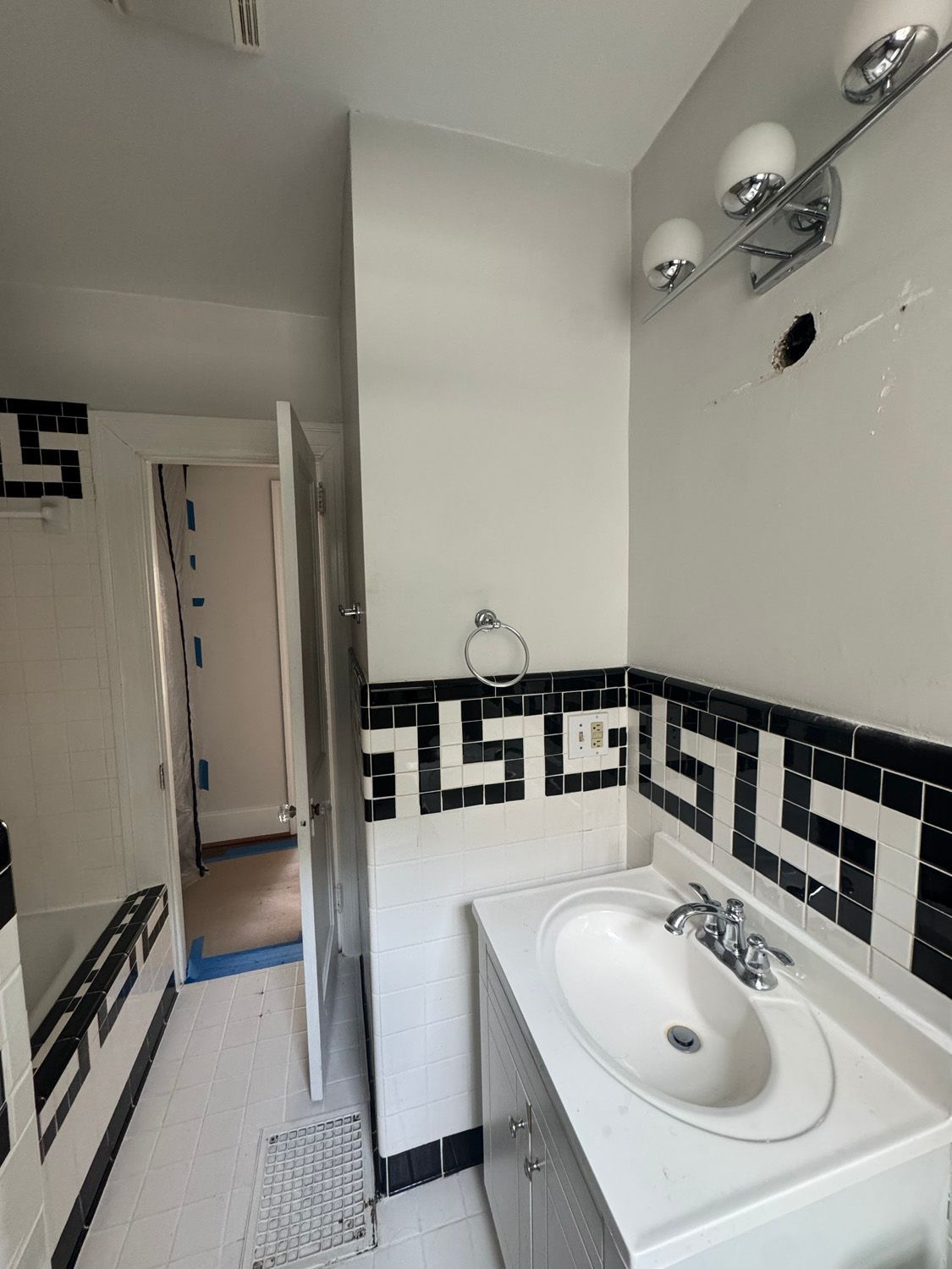 Narrow bathroom with black-and-white tile trim, pedestal sink, wall lights, and open door to a hallway