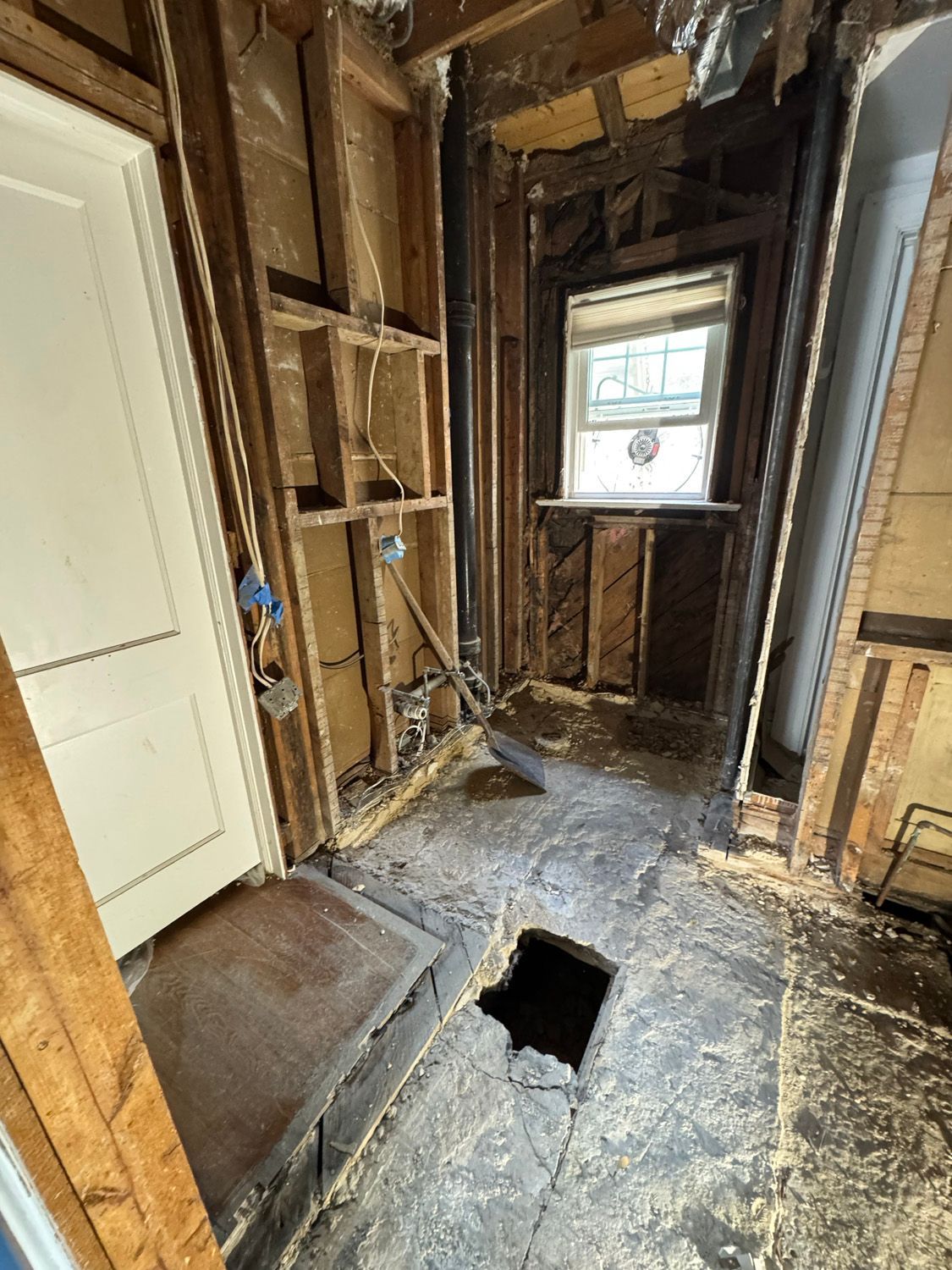 Partially demolished room with exposed studs, debris, and a hole in the floor near a doorway and window
