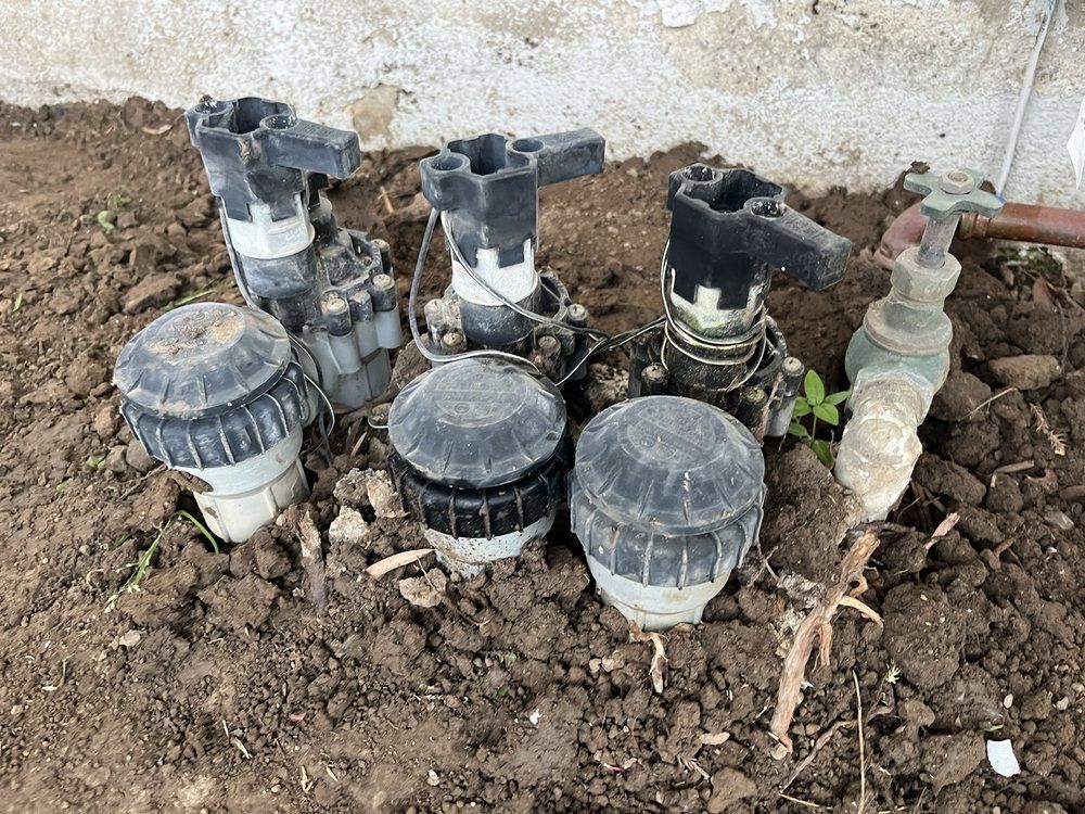 Three black irrigation valve boxes with black tops and a faucet in a dirt garden bed.