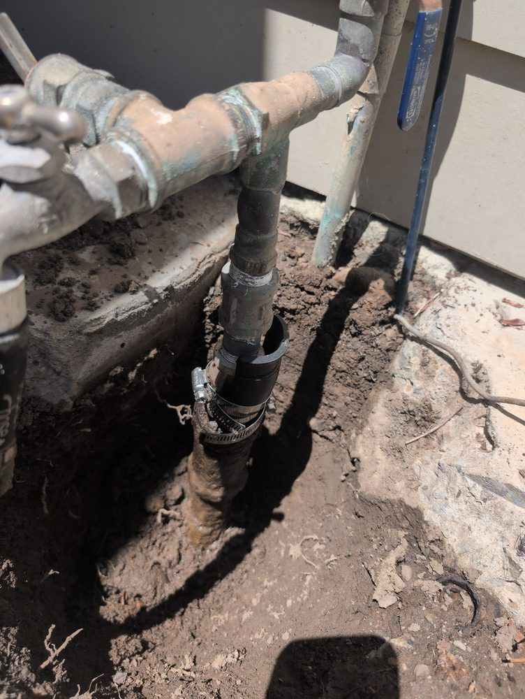 Pipes and fixtures near a wall, buried in dirt with exposed pipe and a gap.