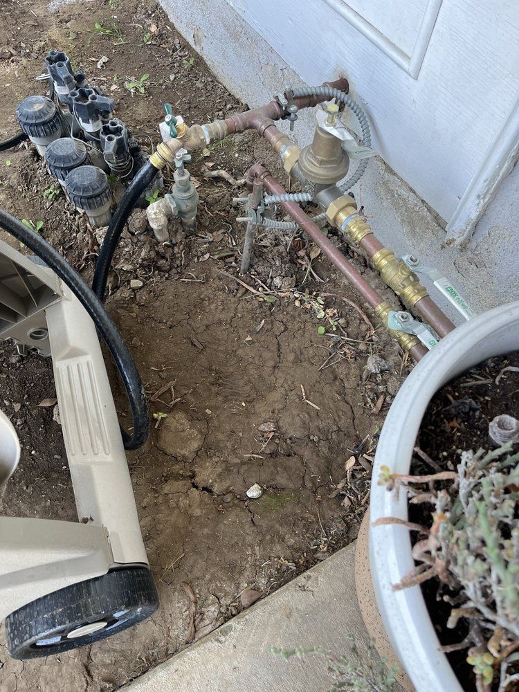 Irrigation system components near a house, including valves, pipes, and sprinklers, on dirt ground.