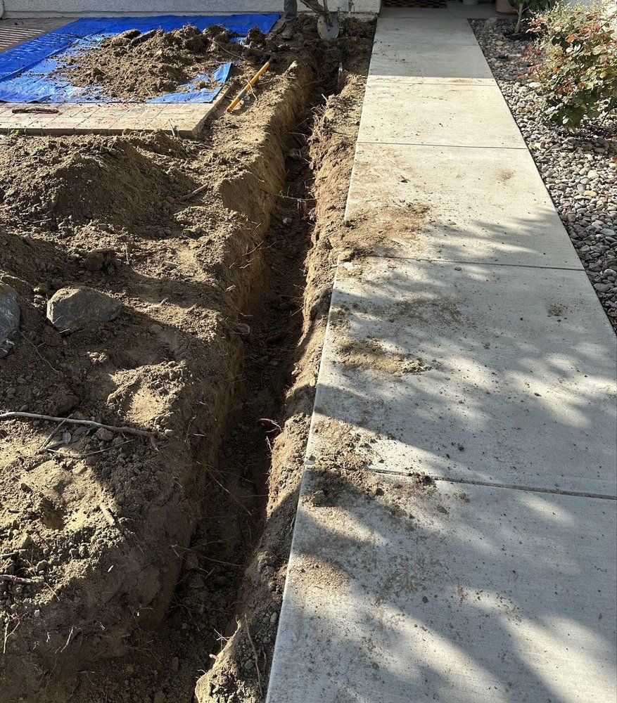 A narrow trench dug alongside a concrete sidewalk, presumably for underground utilities. Earth is on both sides.