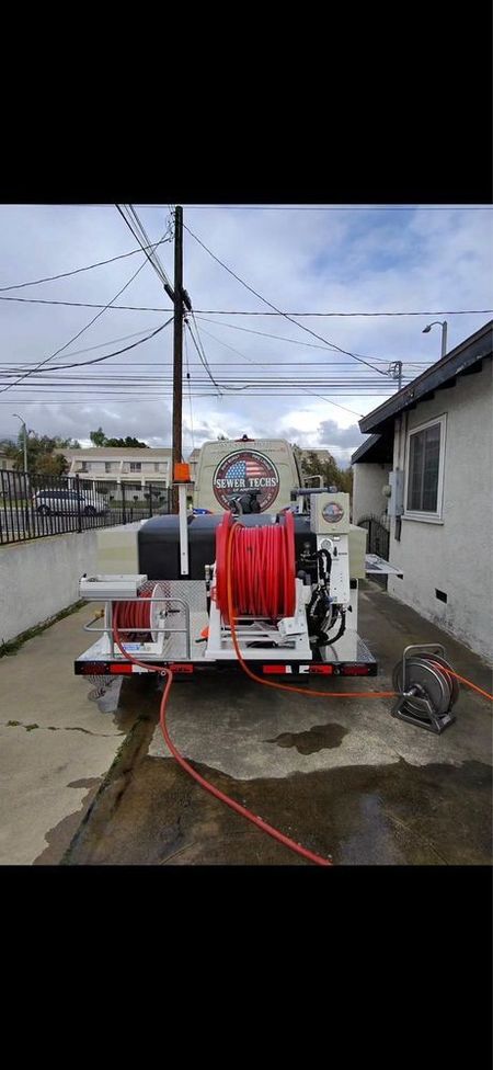 A utility trailer with red hoses and equipment parked between a building and a concrete wall.