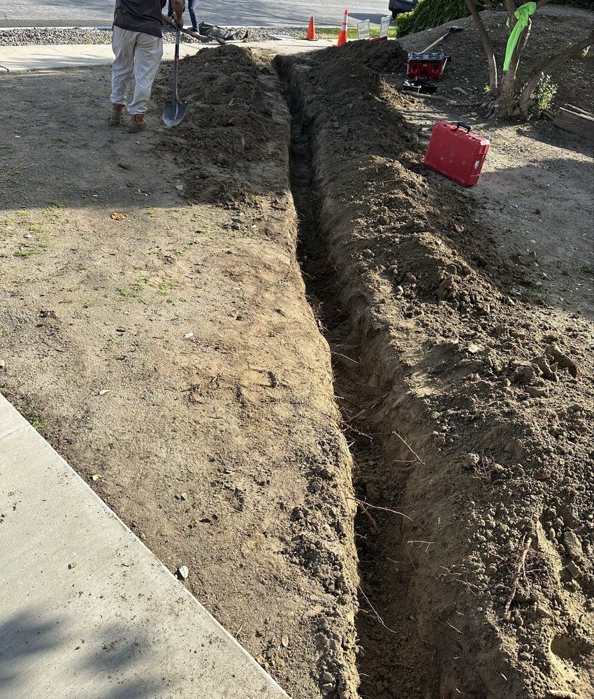 Man digging trench on dirt next to a sidewalk. Orange cones and red toolbox visible.