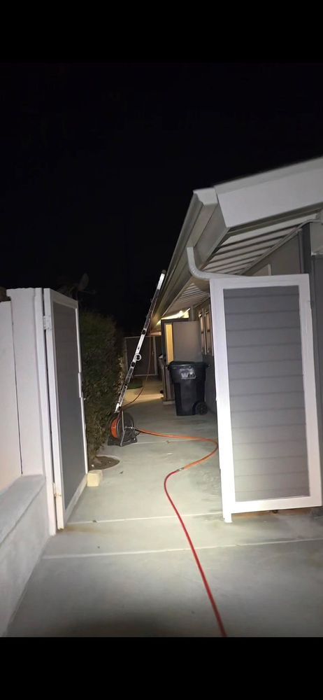 A narrow outdoor space at night with a ladder against a building, a trash can, and a red hose on the ground.