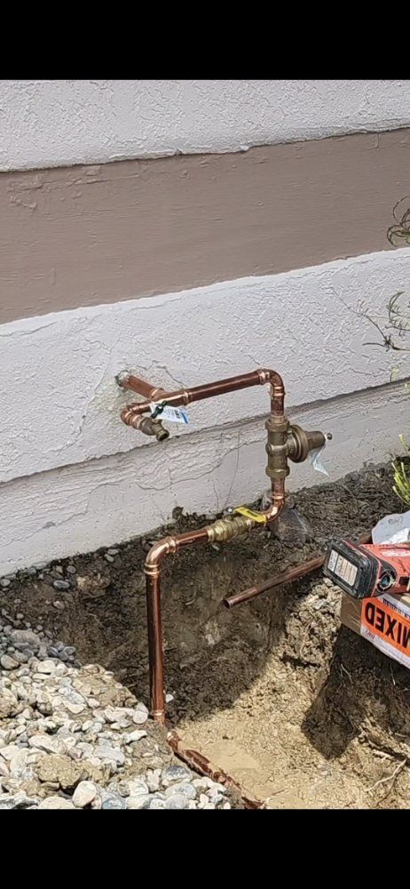 Copper pipes installed near a building's foundation.  A trench holds the lower pipes and fittings.