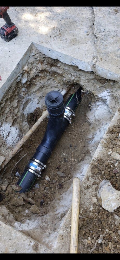 Black pipe with clamps exposed in a dirt pit, alongside a concrete surface and a power drill.