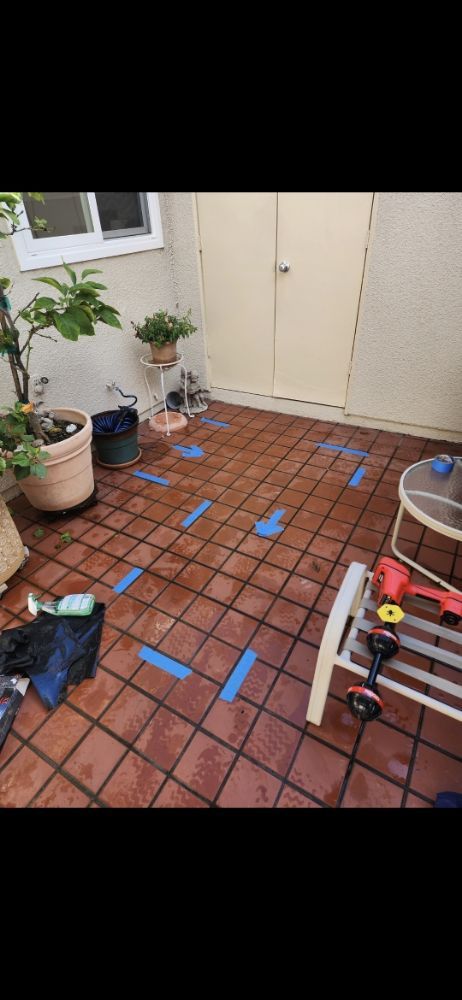 Patio with terracotta tiles marked with blue tape, plants, and tools.