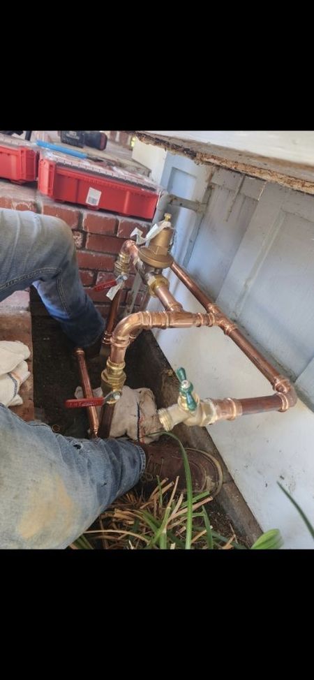 Plumbing work in progress with copper pipes, valves, and a pressure regulator. Person in blue jeans works outdoors.