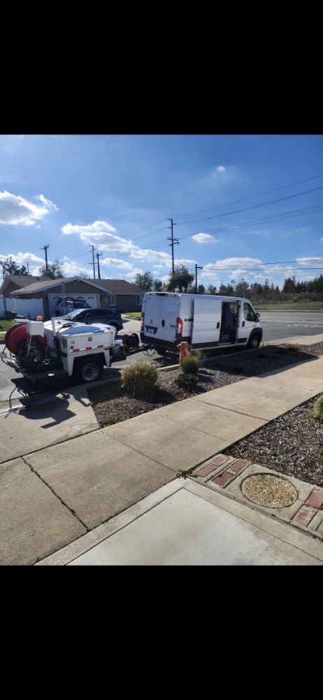 A white utility van parked in front of a house with a trailer; a bright blue sky.
