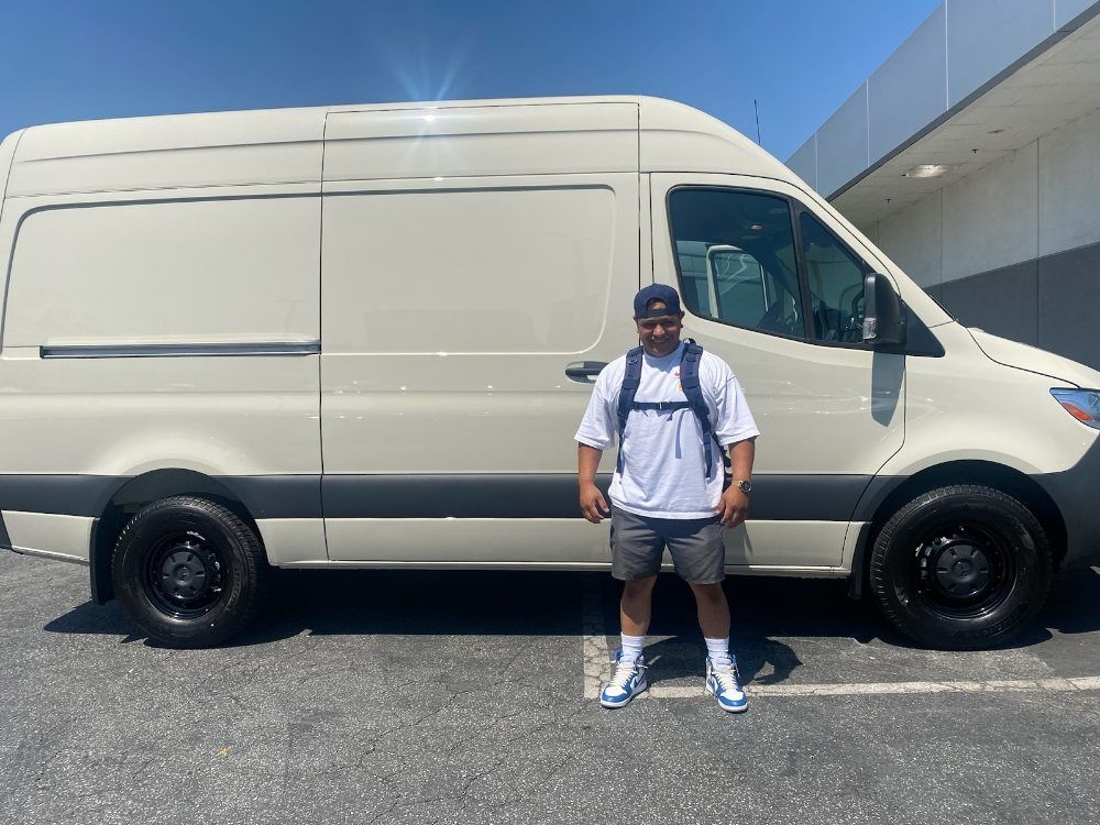 Man standing next to a white van in a parking lot; blue hat, backpack, and shorts.
