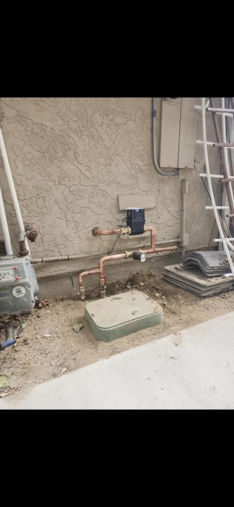 Water meter, copper pipes, and valve near a stucco wall and ground cover.