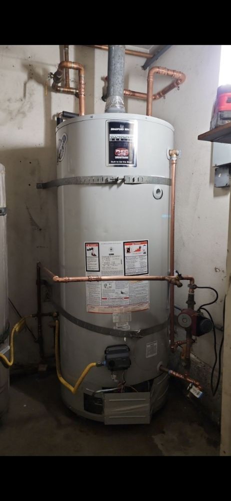 A tall, gray water heater in a basement setting, with copper pipes and yellow wiring visible.