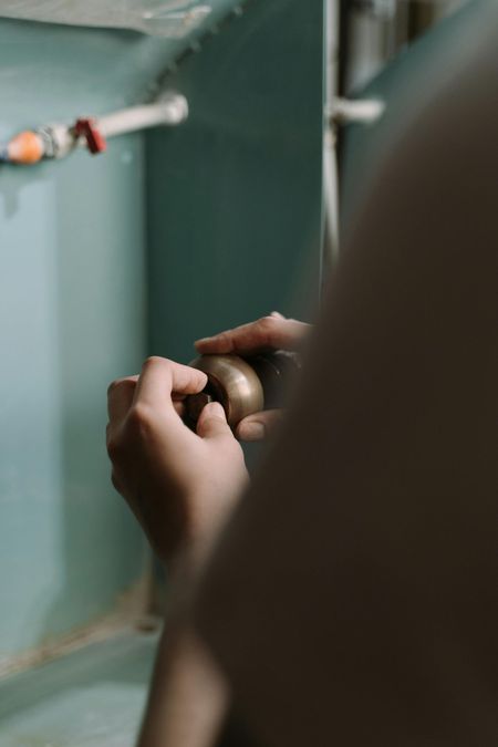 Person holding a small, gold-colored object with hands in a room with a blue wall and pipes.