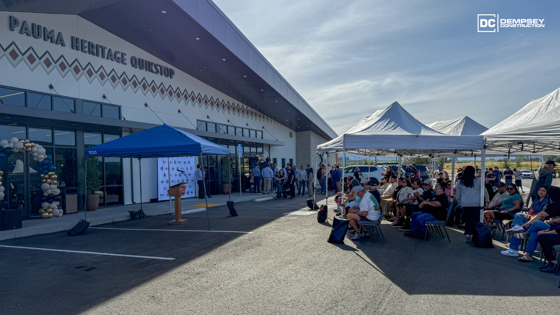 People sit under white event tents at an outdoor gathering in front of the Pauma Heritage Building on a sunny day.