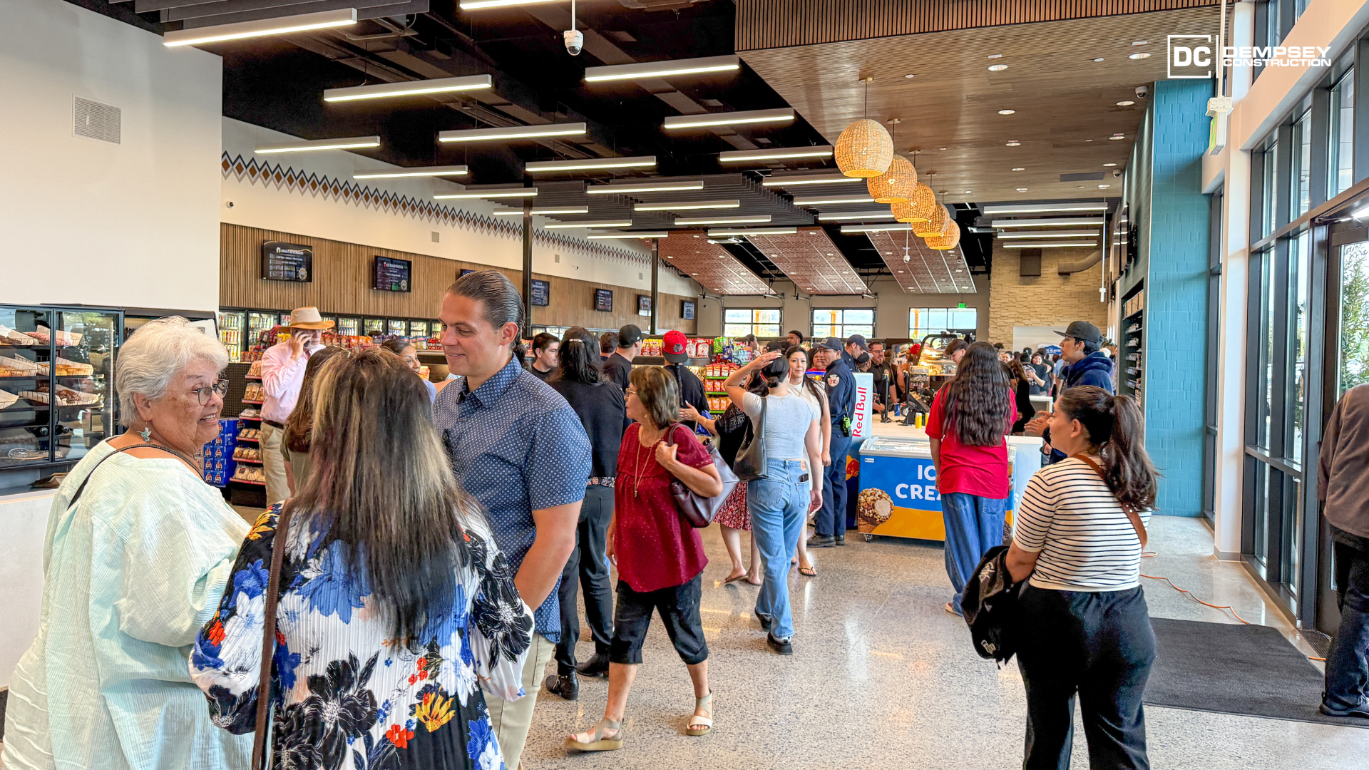Crowded indoor market with people browsing displays, bright overhead lighting, and shelves along the walls.