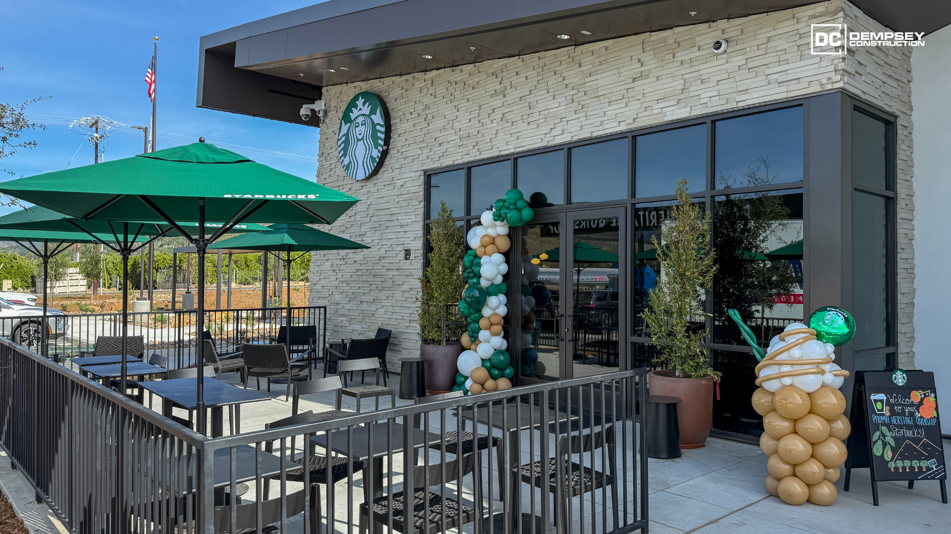 A Starbucks cafe exterior featuring outdoor seating with green umbrellas, a balloon arch, and a large balloon sculpture.