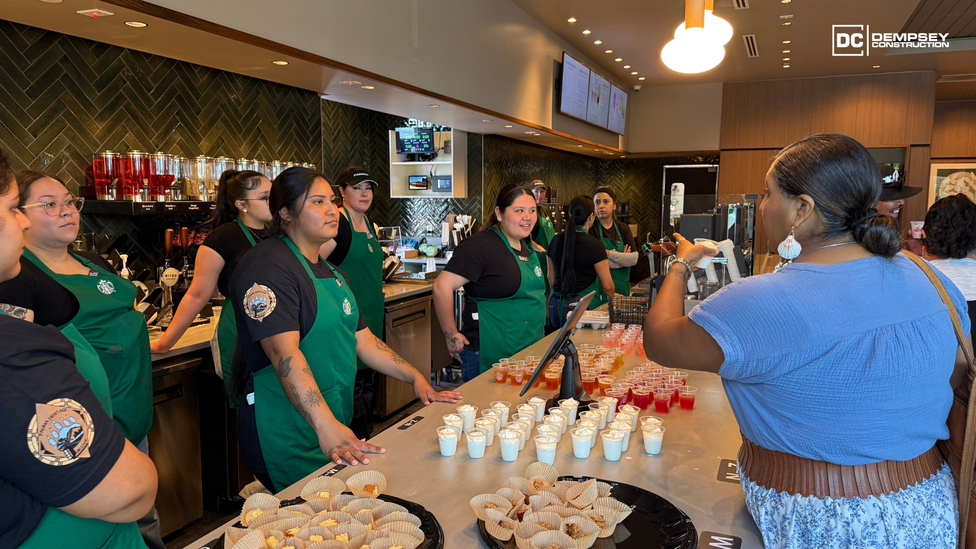 Baristas in green aprons stand behind a counter set with small dessert cups, interacting with customers in a cafe.