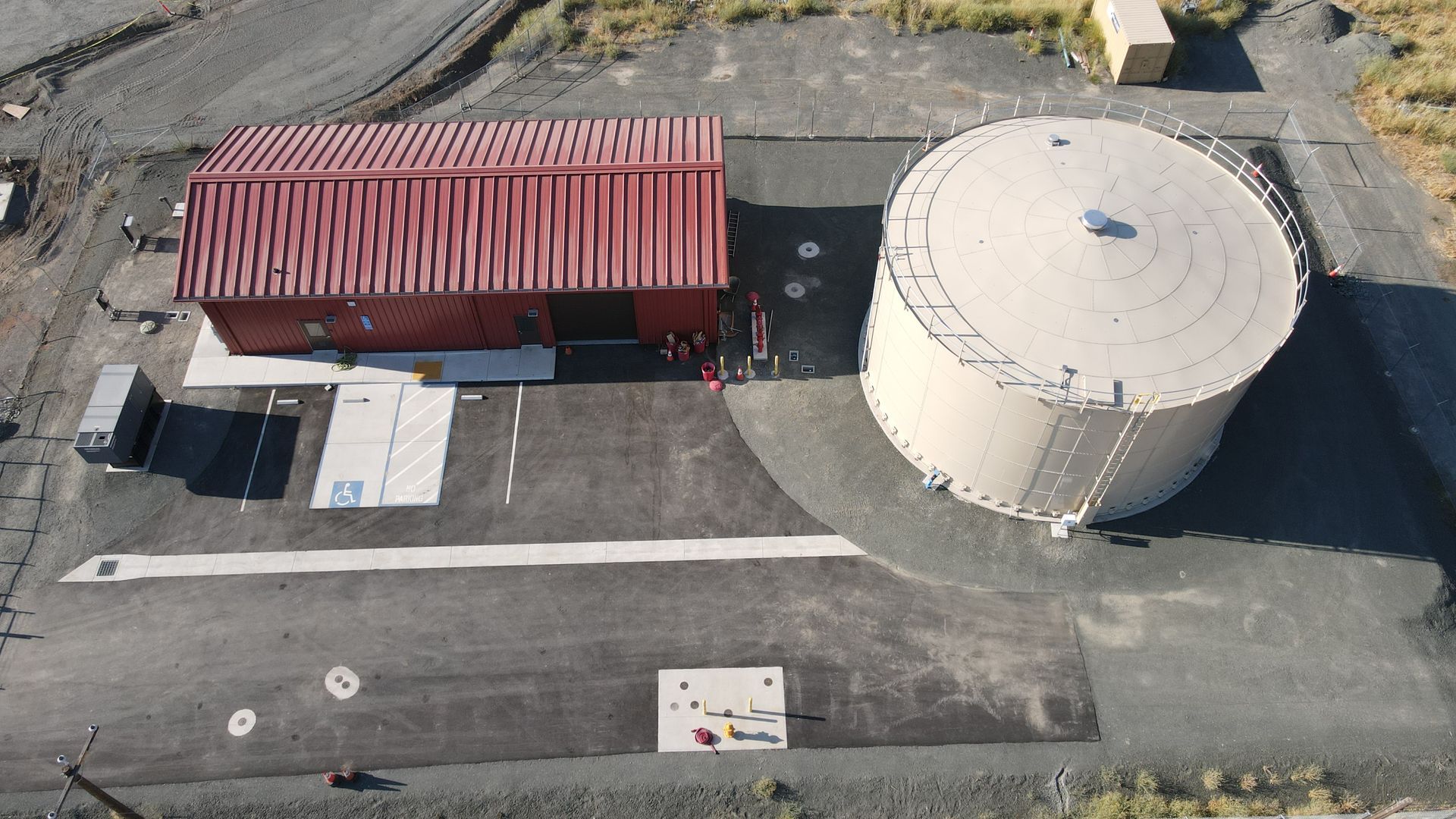Red-roofed building and beige water tank on a paved area surrounded by barren land.