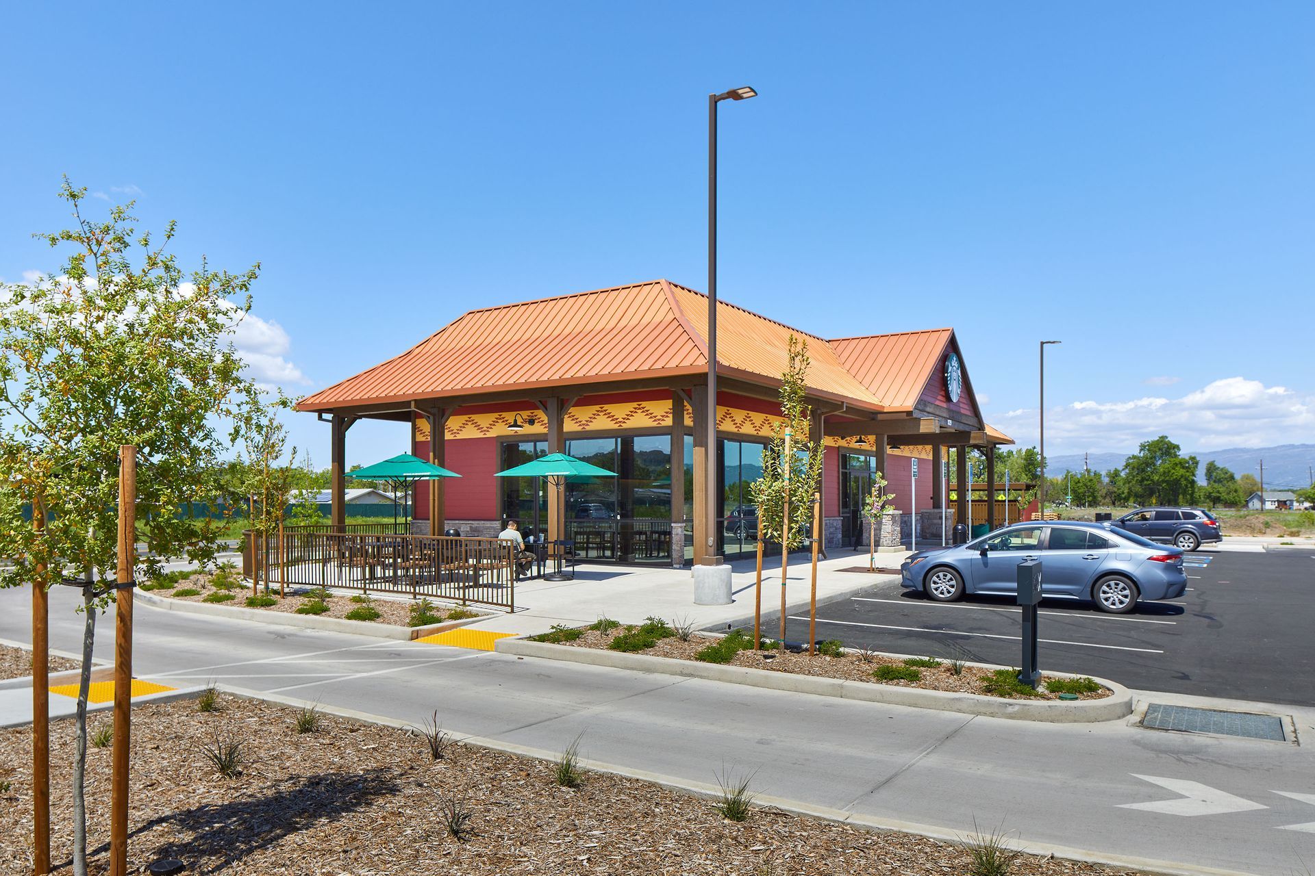 Small pavilion with a red tile roof in a parking lot on a sunny day, with parked cars and young trees.