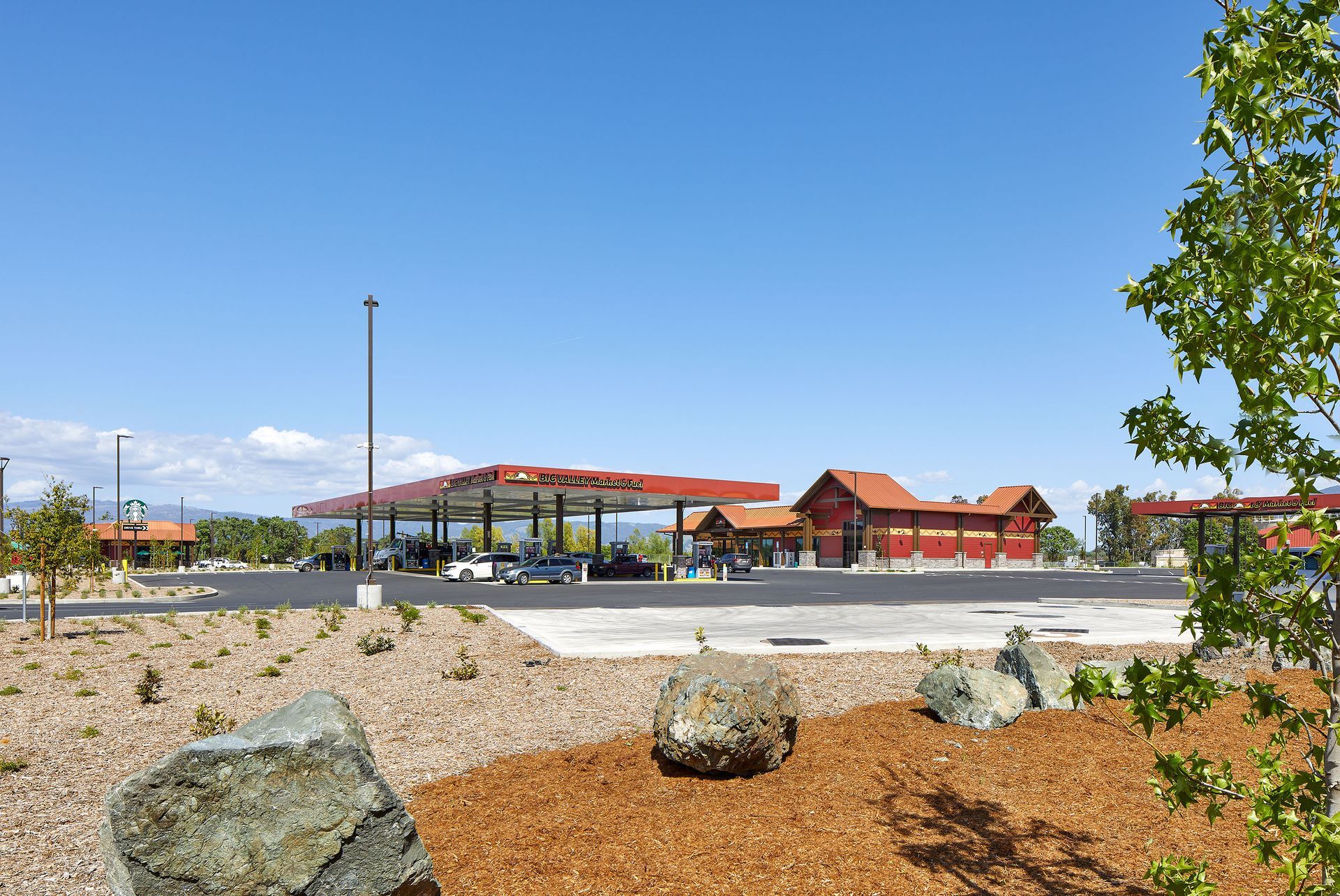 Wide view of a gas station and convenience store under a clear blue sky, with landscaped rocks in front