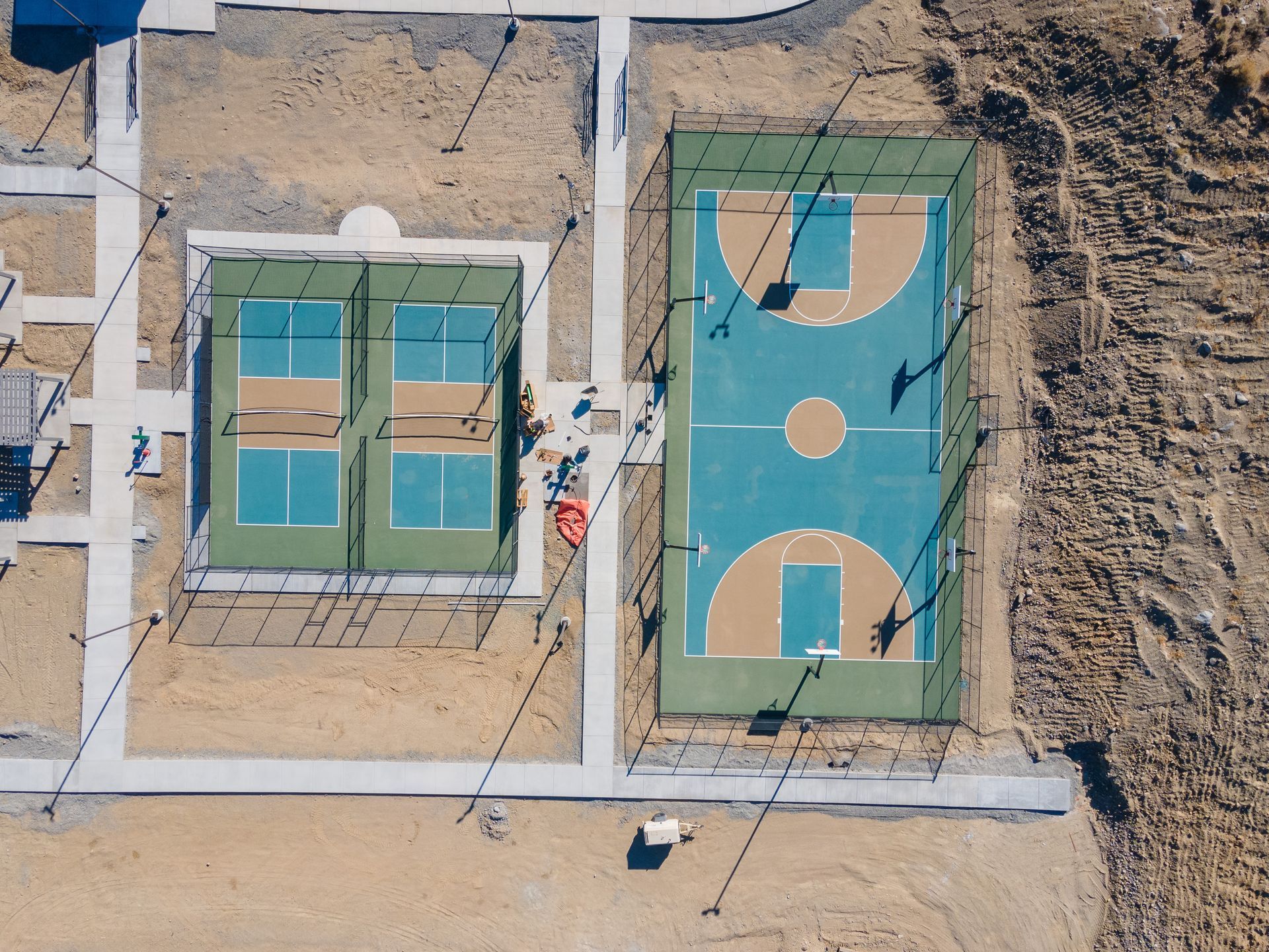 An aerial view of a recreational area featuring two pickleball courts next to a basketball court on a desert landscape.
