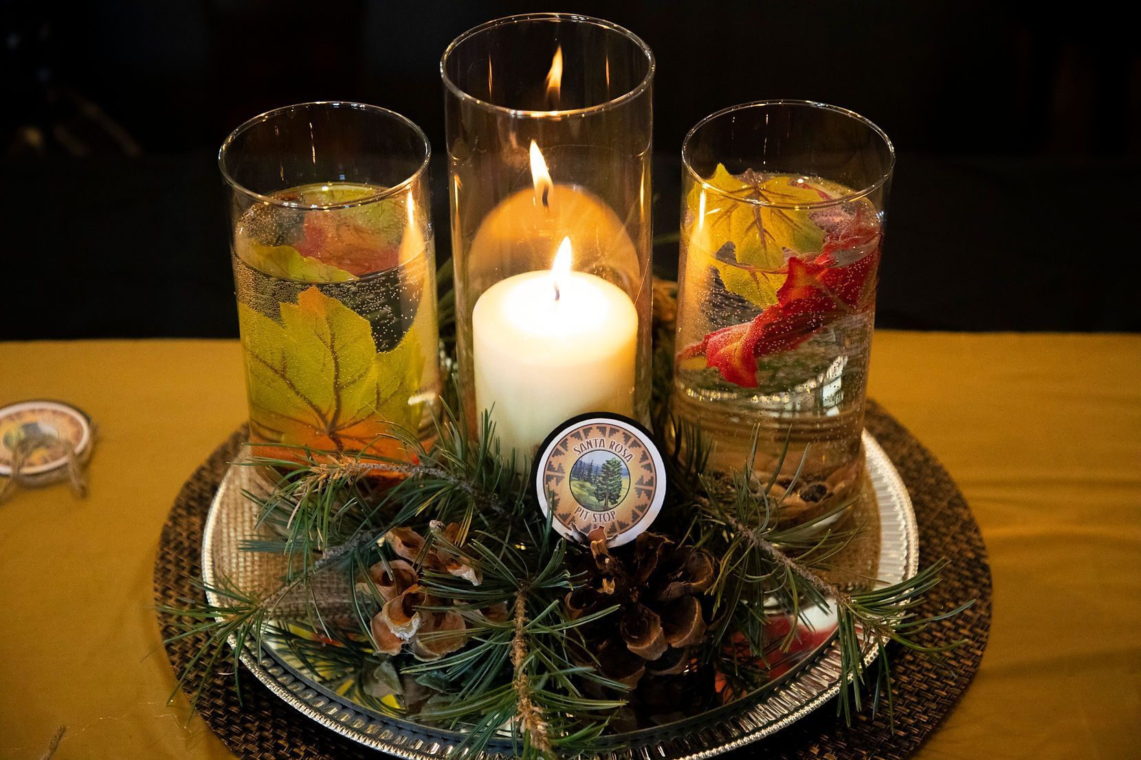 A candle in a glass surrounded by leaves and pine cones on a table.