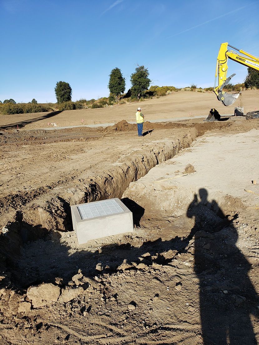 A man is standing in a dirt field with a yellow excavator in the background.
