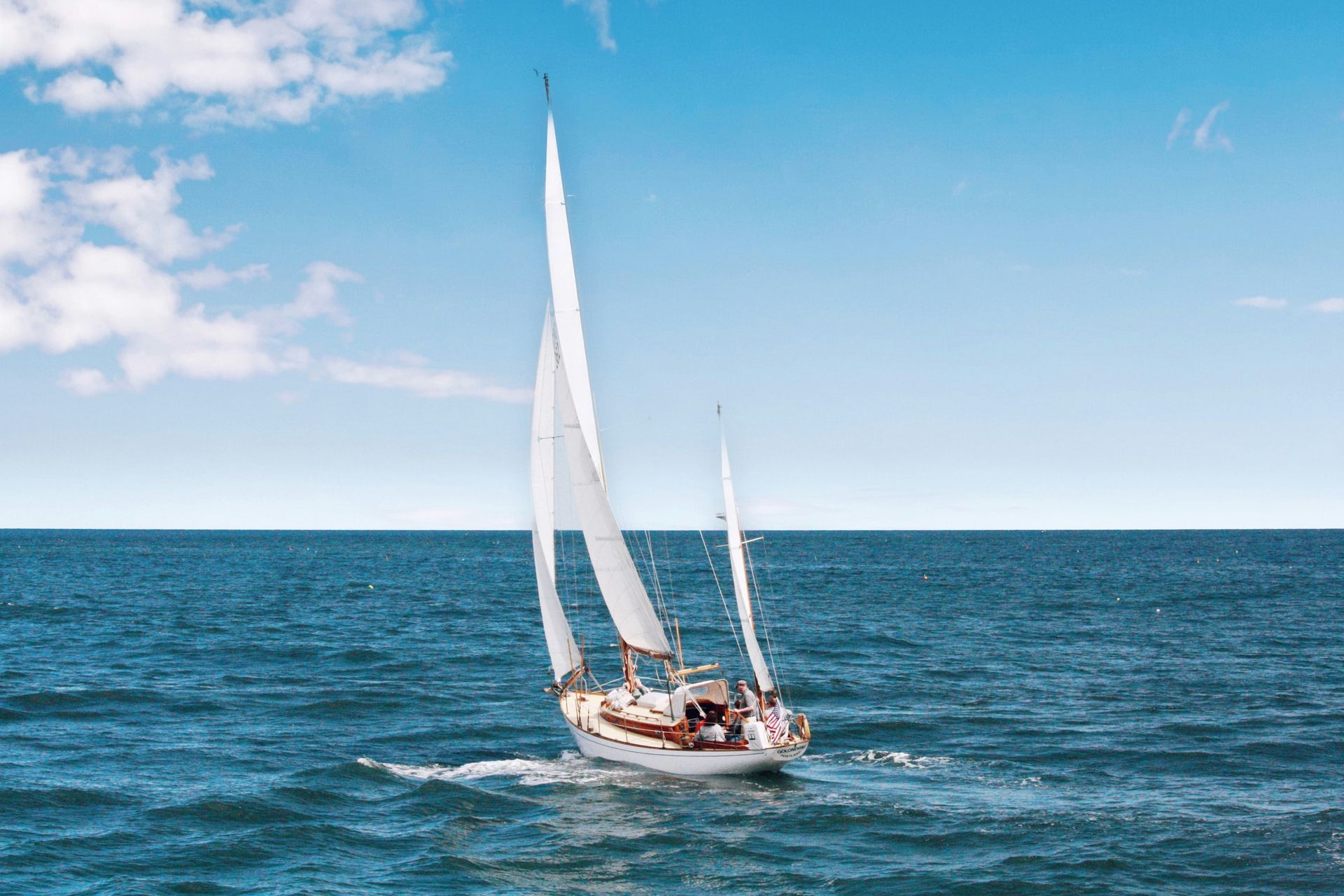 Sailboat on blue ocean with white sails billowing under a partly cloudy sky.