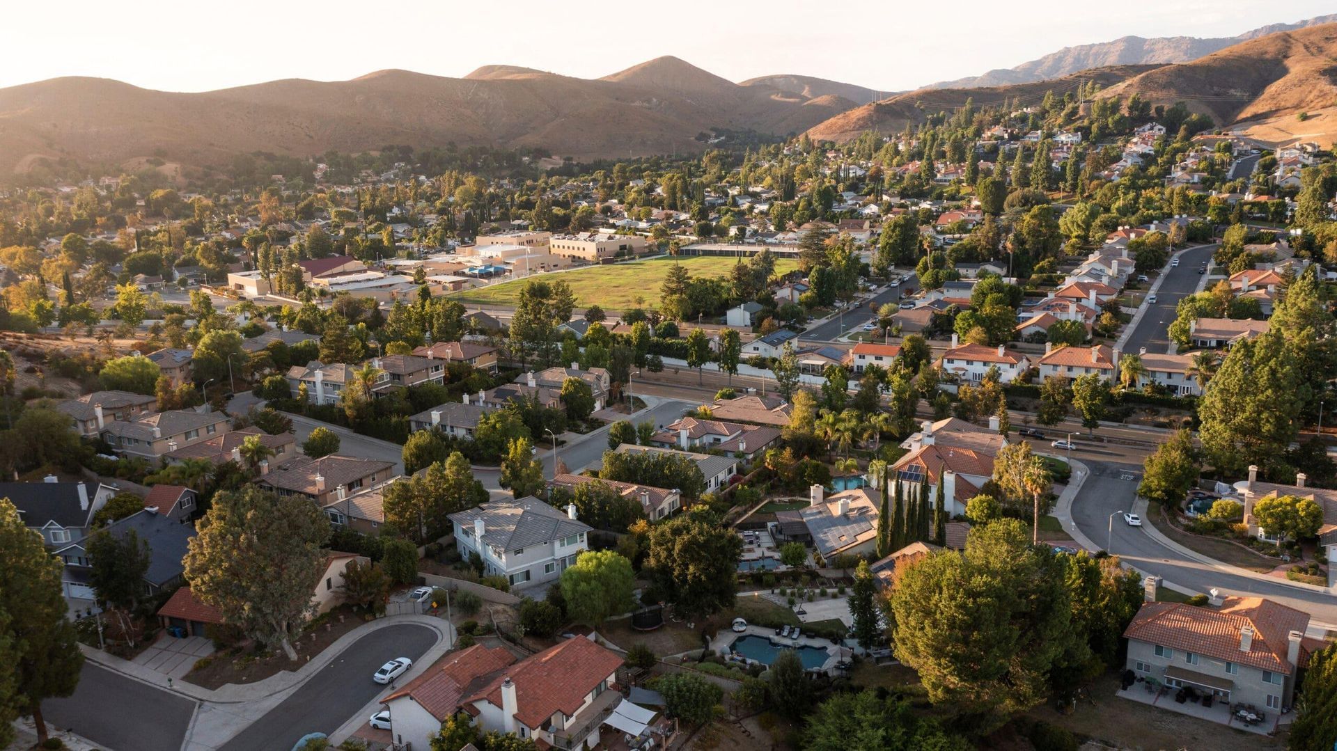 Aerial view of a suburban neighborhood at sunset with houses, trees, winding streets, and hills in the background
